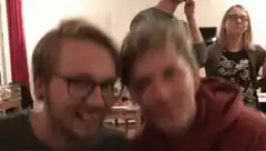 Two men smile at camera while friends celebrate behind