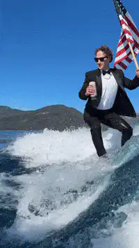 Man in tuxedo surfing with flag and drink