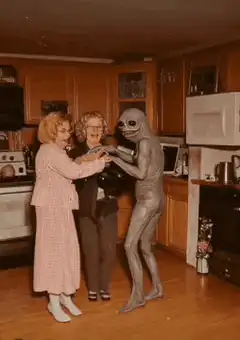Two women dancing with an alien in a kitchen