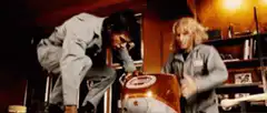 Two men in jumpsuits struggle with a large orange helmet