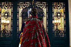 Two guards open ornate doors for a woman in red