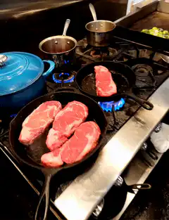 Raw steaks sizzling in cast iron skillets on a gas stove
