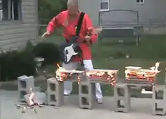 man in red karate uniform plays guitar on burning cinder blocks