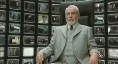 The Director shrugs in front of monitors
