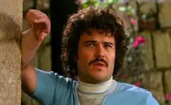 Man with curly hair and mustache leaning on wall